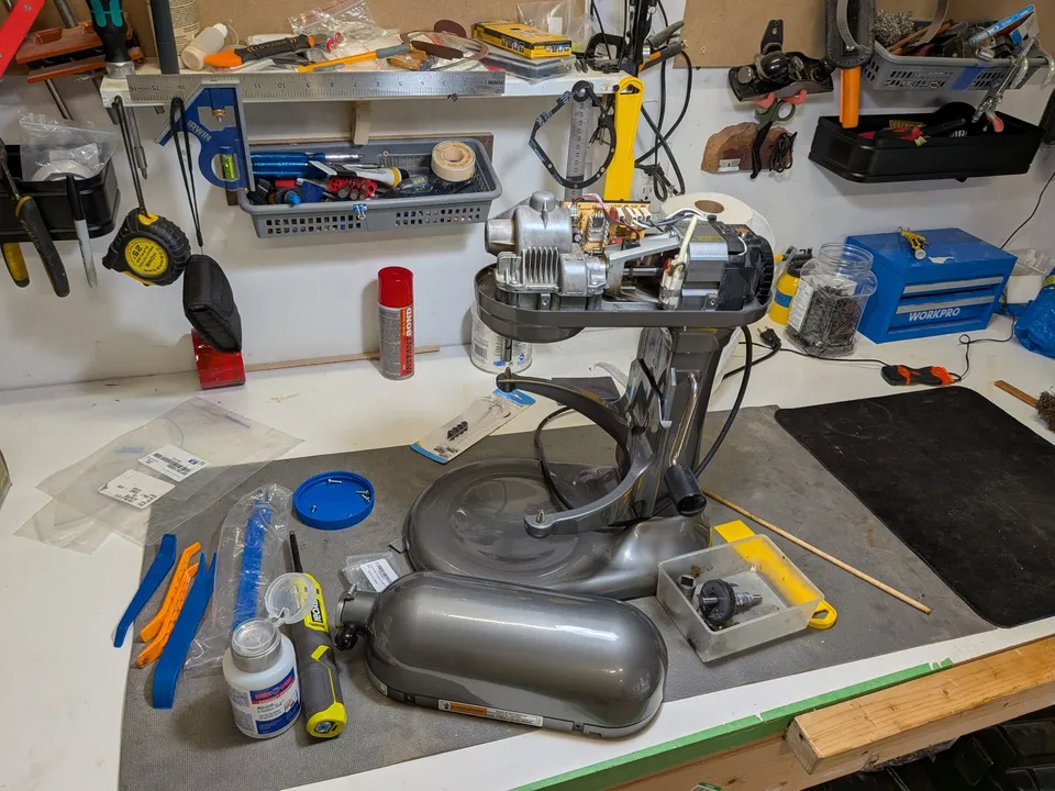 A grey bowl-lift KitchenAid mixer on the MixerFixer workbench with its cover off, showing the gearbox, control board and motor, with the old worm gear in a parts tray beside it